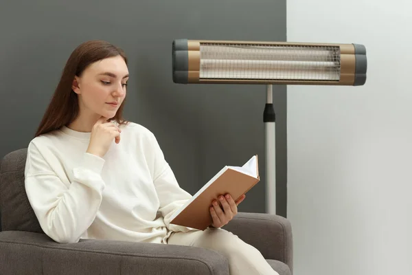 Νεαρή Γυναίκα Διαβάζει Βιβλίο Για Πολυθρόνα Κοντά Ηλεκτρικό Υπέρυθρο Θερμοσίφωνα — Φωτογραφία Αρχείου