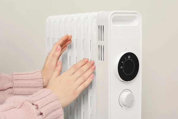 Young woman warming hands near modern electric heater on beige background, closeup