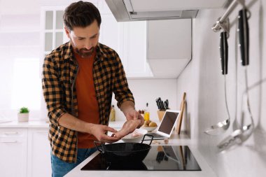Adam mutfakta dizüstü bilgisayarla yemek kursunu izlerken tavuklu filetoyu tavaya koyuyor.