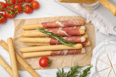 Delicious grissini sticks with prosciutto and snacks on white table, flat lay