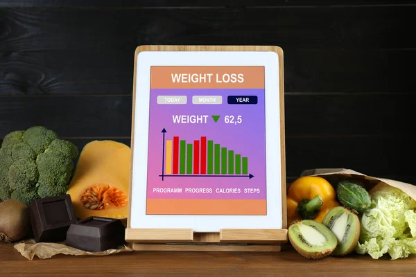 Tablet with weight loss calculator application and food products on wooden table