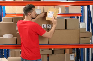 Post office worker holding adhesive paper bag near rack with parcels indoors