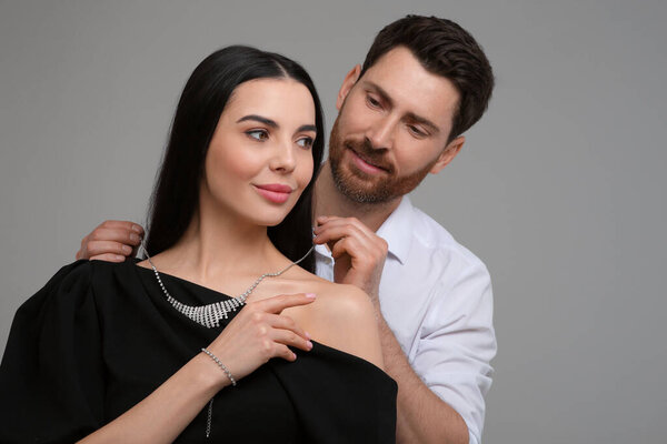 Man putting elegant necklace on beautiful woman against grey background