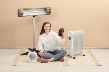 Bej duvarın yanında farklı modern elektrikli ısıtıcıları olan genç bir kadın.