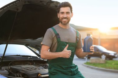 Smiling worker holding blue container of motor oil and showing thumbs up near car outdoors