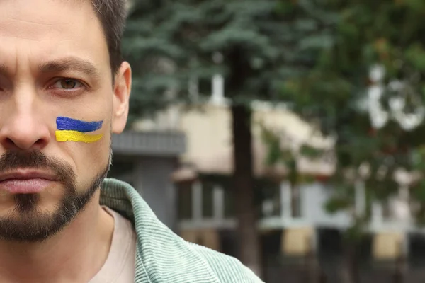 Sad man with drawings of Ukrainian flag on face outdoors, closeup. Space for text