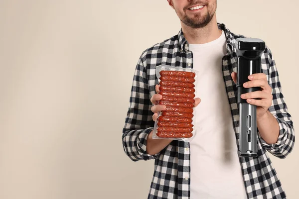 Smiling man holding sous vide cooker and sausages in vacuum pack on beige background, closeup. Space for text