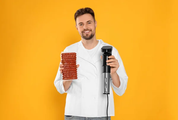 Smiling chef holding sous vide cooker and sausages in vacuum pack on orange background