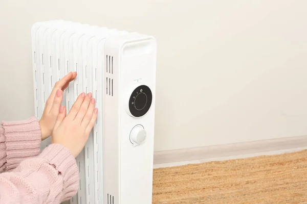 Young woman warming hands near modern electric heater against beige background indoors, closeup