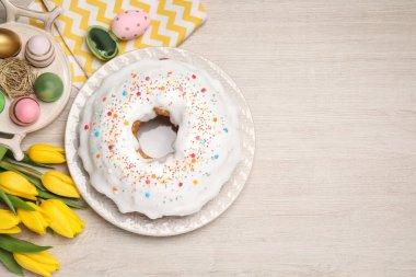 Easter cake with sprinkles, painted eggs and tulips on white wooden table, flat lay. Space for text