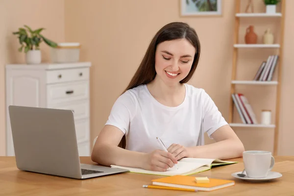 快乐的年轻女子在屋里木桌边的笔记本上写字 — 图库照片