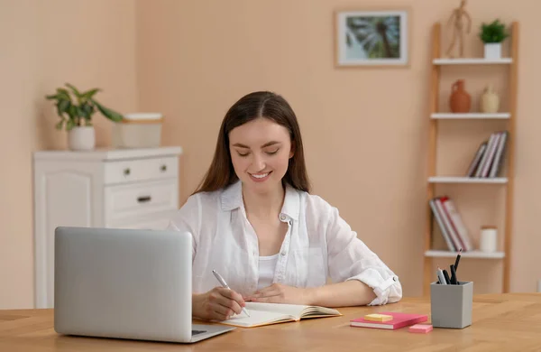 快乐的年轻女子在笔记本上写字 同时在室内木制桌旁的笔记本电脑上工作 — 图库照片