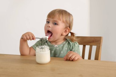 Cute little child eating tasty yogurt with spoon at wooden table indoors clipart