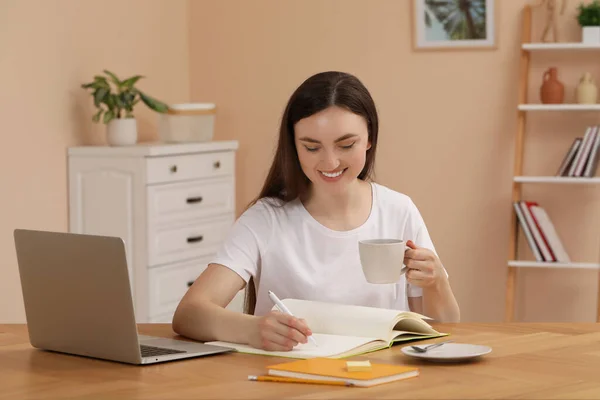 年轻的女人端着杯子喝咖啡 在屋里的木制桌上用笔记本写字 — 图库照片