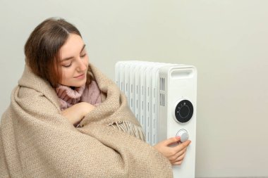 Bej duvarın yanındaki modern elektrikli ısıtıcının ısısını ayarlayan genç bir kadın.