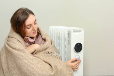 Young woman adjusting temperature on modern electric heater near beige wall clipart