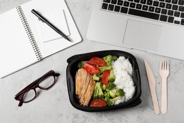 Lezzetli yemek, dizüstü bilgisayar, plastik çatal bıçak takımı ve açık gri masa üzerinde düz bir defter. İş yemeği.