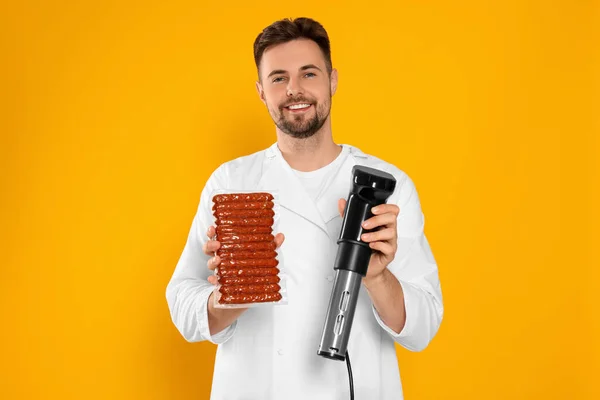 Smiling chef holding sous vide cooker and sausages in vacuum pack on orange background