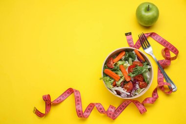 Ölçüm bandı, sebze salatası ve sarı arka planda elma, mesaj için boş alan. Kilo verme konsepti