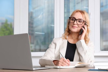 Bayan patron ofisteki bilgisayarın yanında akıllı telefondan konuşuyor. Başarılı bir iş kadını.