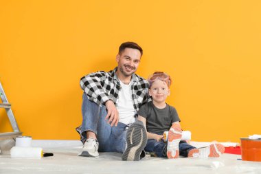 Turuncu duvarın yanında tamir aletleri olan baba oğul.