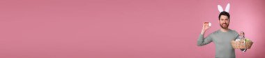 Tavşan kulaklı mutlu adam pembe arka planda paskalya yumurtası dolu sepeti tutuyor, mesaj için yer var. Pankart tasarımı