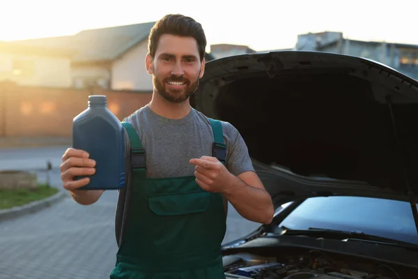 Smiling worker pointing at blue container of motor oil near car outdoors