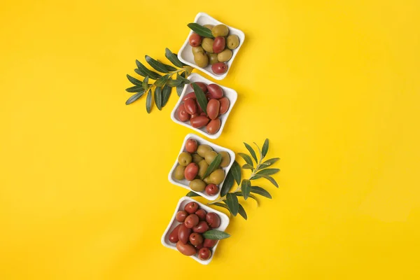 Different fresh olives and green leaves on yellow background, flat lay