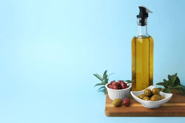 Bottle of oil, olives and tree twigs on light blue background, space for text