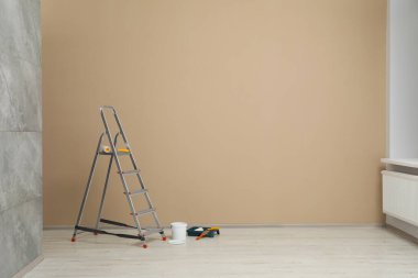 Metallic folding ladder and painting tools near beige wall indoors, space for text clipart