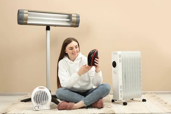 ベージュの壁の近くに異なる近代的な電気ヒーターを持つ若い女性屋内 — ストック写真