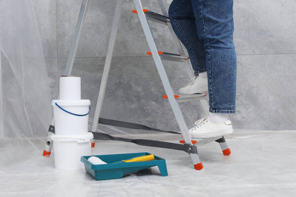 Woman climbing metallic folding ladder, tray with roller and buckets of paint indoors, closeup