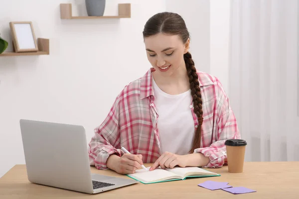 快乐的年轻女子在笔记本上写字 同时在室内木制桌旁的笔记本电脑上工作 — 图库照片