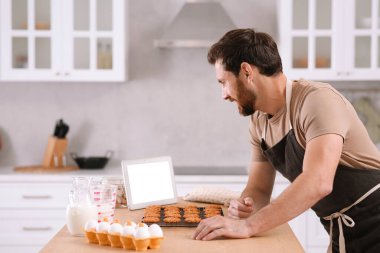 Yeni pişmiş kurabiyeli bir adam mutfakta tabletten çevrimiçi yemek kursunu izliyor.