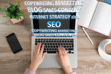 Arama motoru optimizasyonu (SEO). Metin yazarı, ahşap masada dizüstü bilgisayarda çalışıyor. Bilgisayardan pek çok kelime geliyor