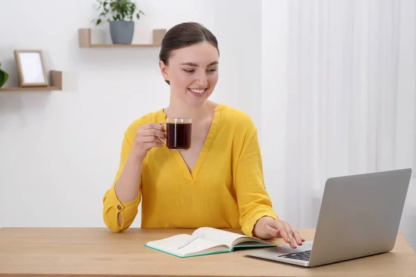 快乐的年轻女人 带着一杯咖啡和笔记本 在屋里的木制桌子边工作 — 图库照片