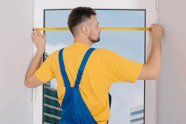 Worker in uniform measuring window with tape indoors. Roller blinds installation