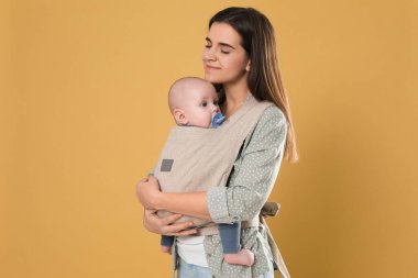 Mother holding her child in sling (baby carrier) on beige background clipart