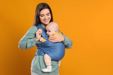 Anne turuncu arka planda çocuğunu askıda (bebek taşıyıcısı) tutuyor. Metin için boşluk