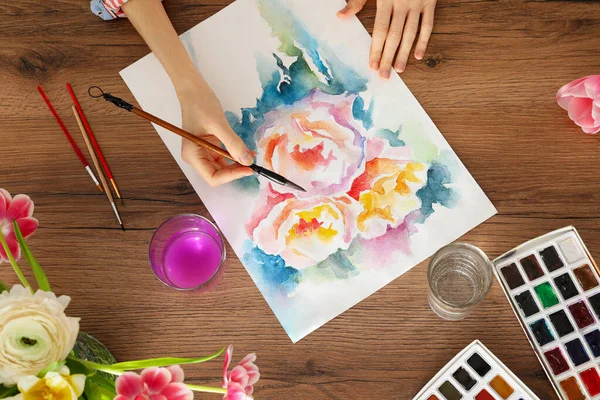 Woman painting flowers with watercolor at wooden table, top view. Creative artwork