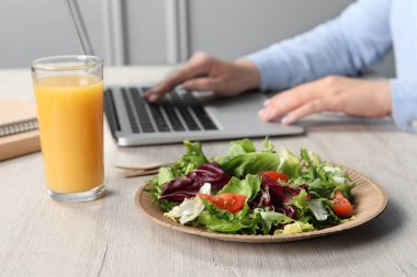 Beyaz ahşap masada dizüstü bilgisayarla çalışan bir ofis çalışanı bir bardak meyve suyu ile taze sebze salatasına odaklanıyor. İş yemeği.