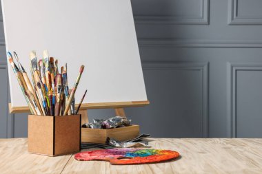 Boş tuvali ve gri duvarın yanındaki ahşap masada farklı sanat malzemeleri olan Easel. Metin için boşluk
