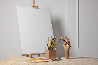 Boş tuval, el modeli ve beyaz duvarın yanındaki ahşap masada farklı sanat malzemeleri.