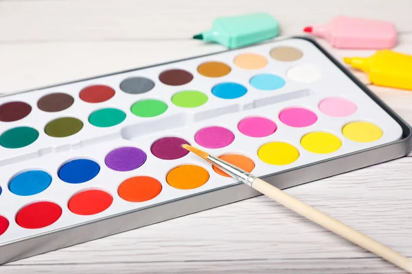 Watercolor palette with brush and colorful markers on white wooden table, closeup
