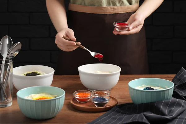 Woman adding red food coloring into bowl at wooden table, closeup