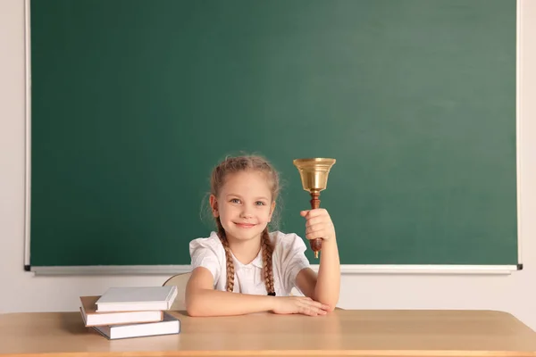Pupil with school bell sitting at desk near chalkboard in classroom, space for text