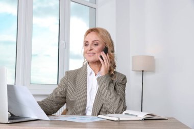 Lady boss with papers talking on smartphone at desk in office. Successful businesswoman