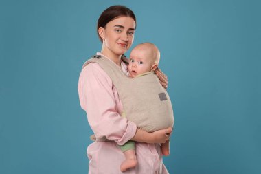 Anne açık mavi arka planda çocuğunu askıda (bebek taşıyıcısı) tutuyor