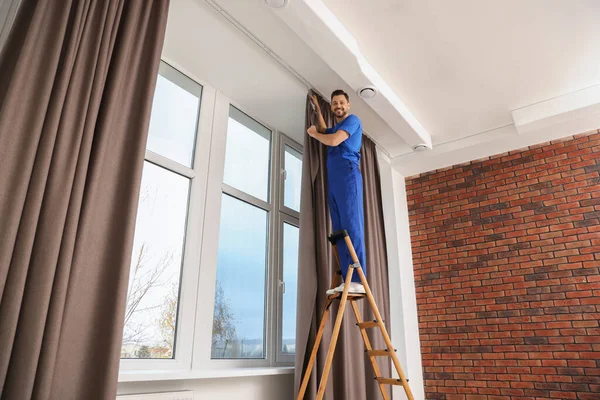Worker in uniform hanging window curtain indoors, low angle view