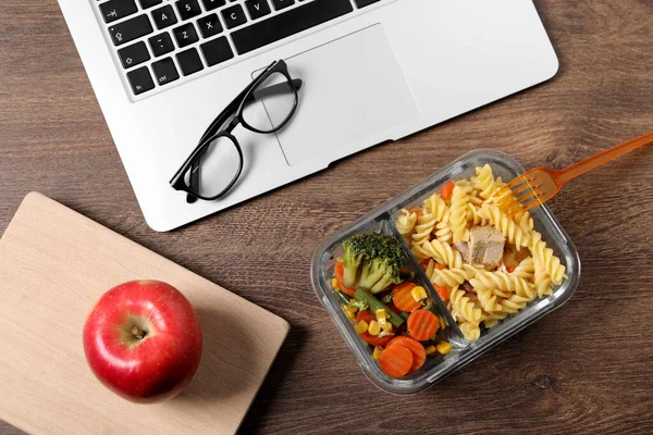 stock image Container of tasty food, laptop, apple, book and glasses on wooden table, flat lay. Business lunch